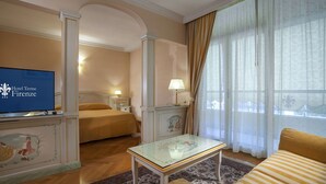 Habitación de hotel con cama, televisión mostrando 'Hotel Termé Firenze', una mesita, un sofá y una ventana con cortinas.