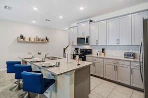 A modern kitchen with white cabinets, a central island with bar stools, a microwave, a toaster, and a refrigerator. - Disney Themes: Resort 5BR Townhouse w Private Pool (Kissimmee)