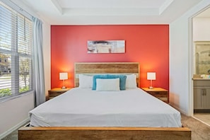 A hotel room with a large bed, two bedside tables with lamps, a red accent wall, and a window with blinds. - Disney Themes: Resort 5BR Townhouse w Private Pool (Kissimmee)