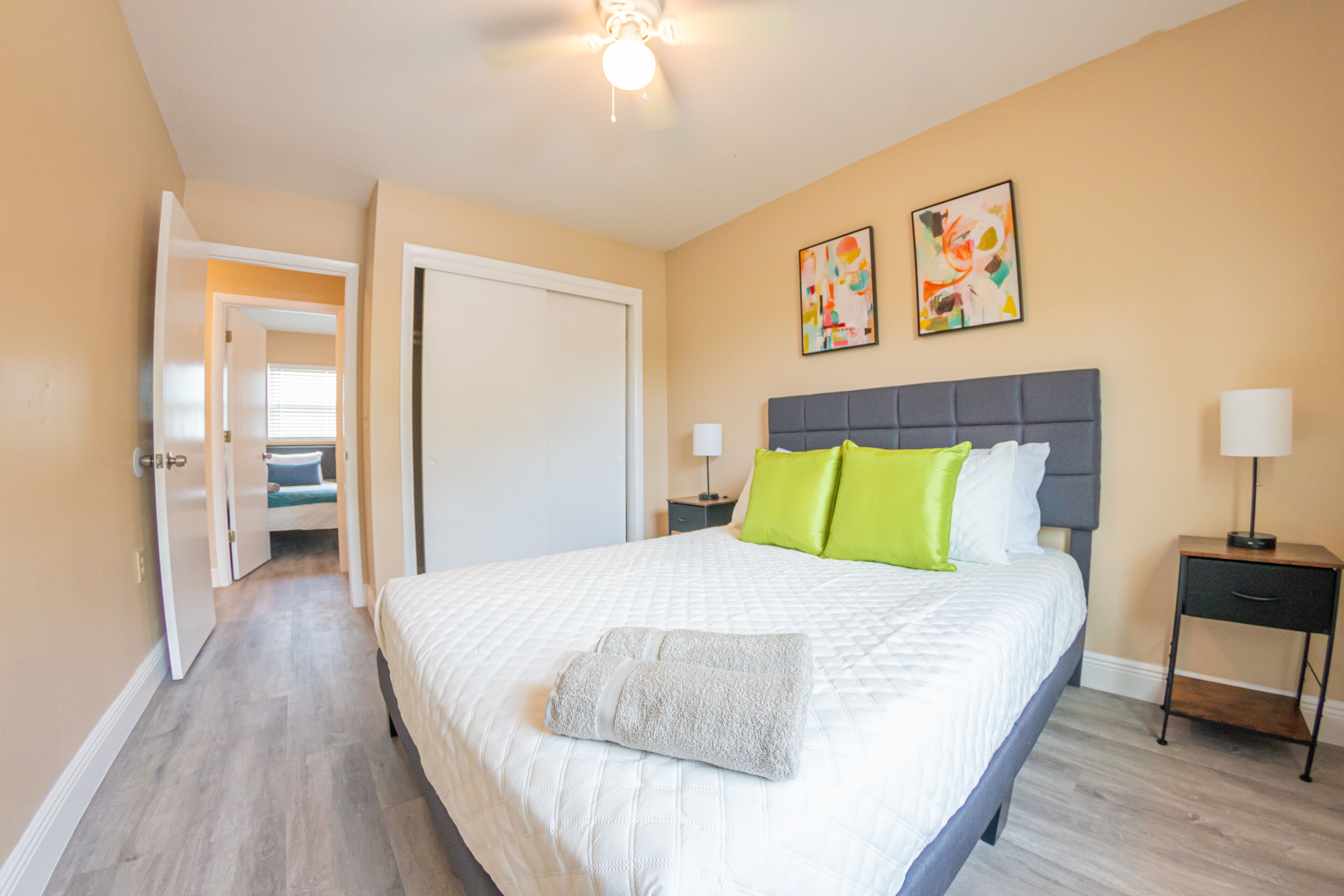 Ein Hotelzimmer mit einem großen Bett, zwei Kissen, einem Nachttisch, einem Deckenventilator und Bildern an der Wand.