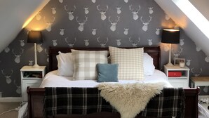 Un dormitorio con techo inclinado, una cama con muchos cojines, dos mesitas de noche con lámparas, y un papel tapiz estampado con motivos de venados.