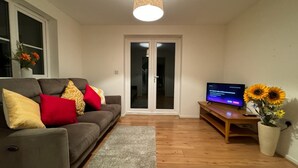 En stue med en grå sofa, røde og gule puder, et træbord og et fladskærms-tv, samt en blomsterdekoration.