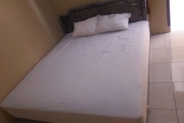 Una cama individual con cabecera de madera y ropa de cama blanca en una habitación con paredes color beige y suelo de baldosas.