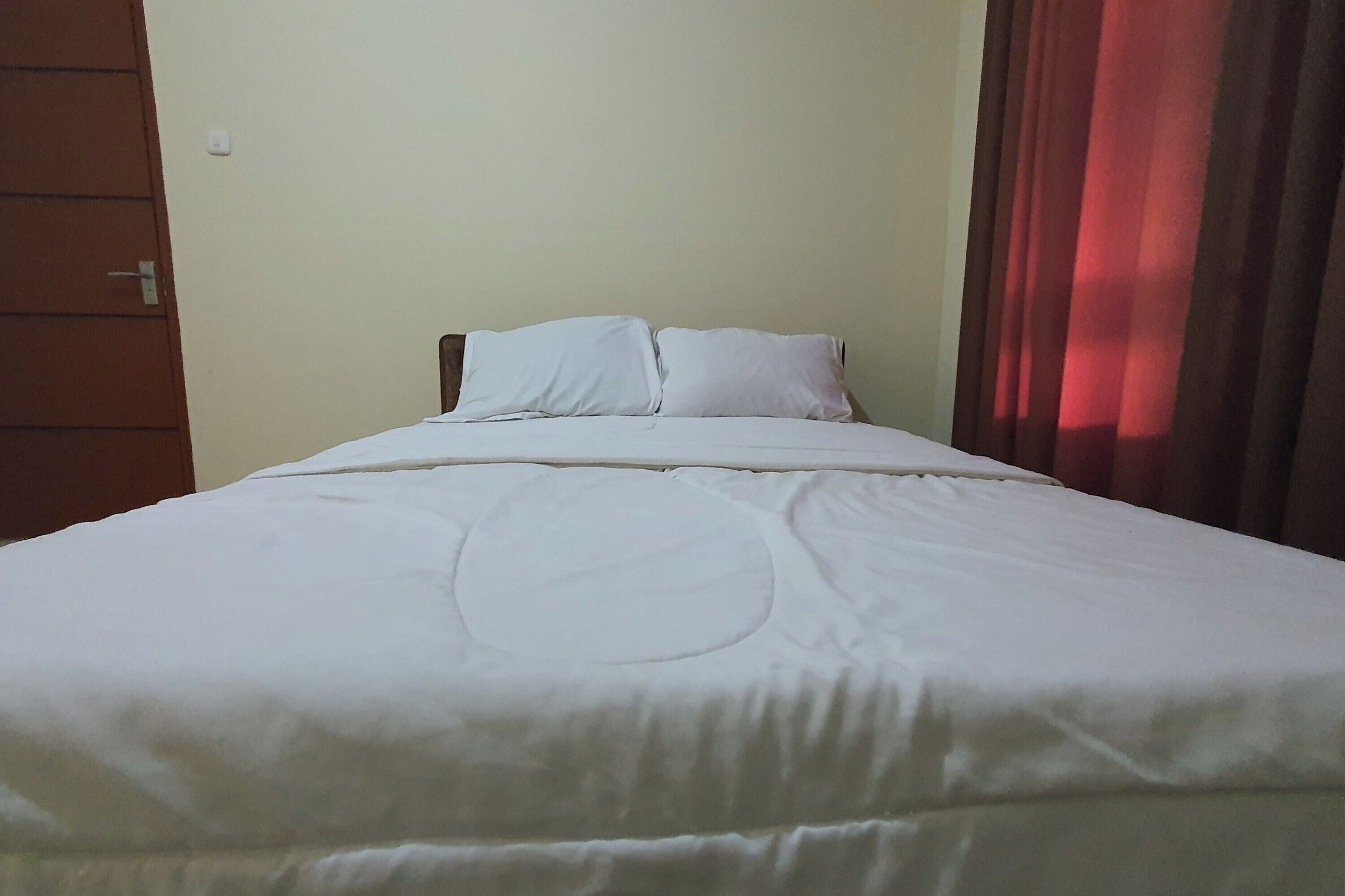 A neatly made bed with a white comforter and pillows, a wooden headboard, and a red curtain partially drawn to the side.
