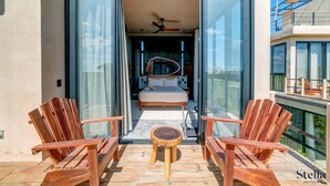 An open balcony with wooden chairs and a small table, leading to a bedroom with a bed and a ceiling fan.