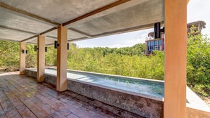A covered outdoor swimming pool with a wooden deck, surrounded by lush greenery and a modern building in the background.