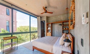A spacious bedroom with a large bed, a ceiling fan, a wooden wardrobe, and a view of greenery outside. - Jungle Studios on Beach Road with Pool  (Tulum)