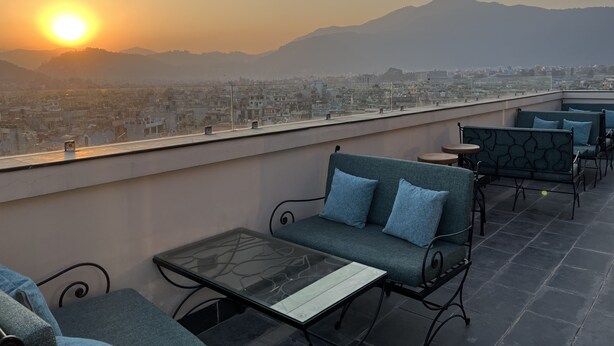 A rooftop terrace with seating and a glass table, overlooking a cityscape and mountains during sunset. - Hotel Kesu Home (Kathmandu)