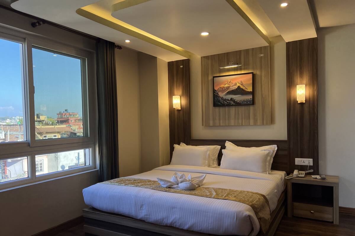 A hotel room with a bed, a nightstand, a wall-mounted lamp, a painting, and a window with curtains.