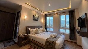 A hotel room with a large bed, a bedside table with a phone, a painting on the wall, and a view of the mountains through the window. - Hotel Kesu Home (Kathmandu)