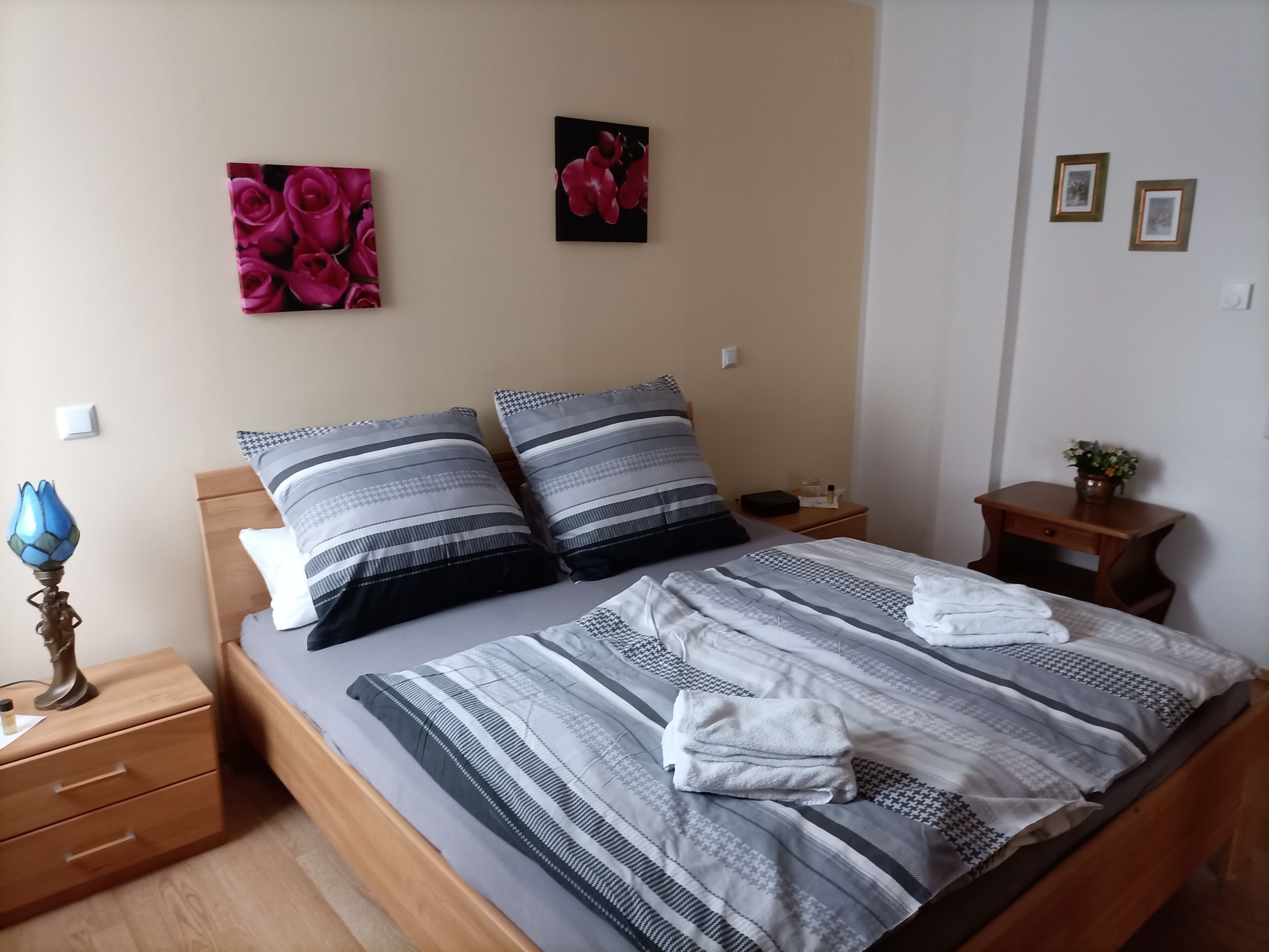 A neatly made bed with striped bedding, two pillows, and a folded towel. There are two bedside tables, one with a lamp and the other with a small plant.