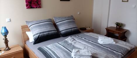 A neatly made bed with striped bedding, two pillows, and a folded towel. There are two bedside tables, one with a lamp and the other with a small plant.