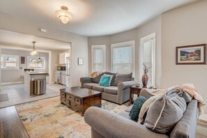 A spacious living room with a grey sofa, a coffee table, and a rug. There is a kitchen area in the background with a refrigerator and a sink. - Hotel Heritage | Historic Sites | Dog Friendly/BBQ (Pueblo)