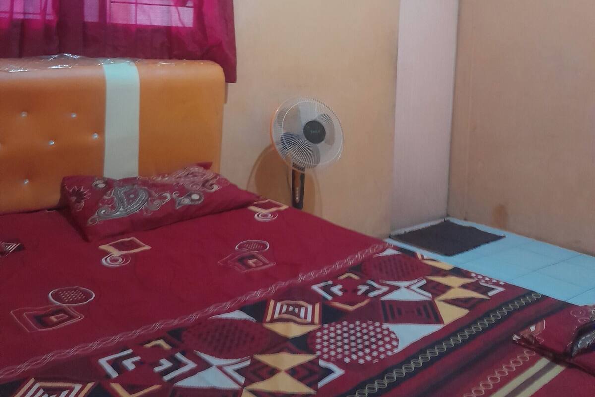 A bedroom with a bed, a standing fan, and a window with curtains.