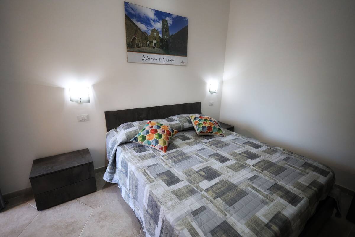 A bedroom with a bed, two wall-mounted lights, a nightstand, a bench, and a welcome sign.