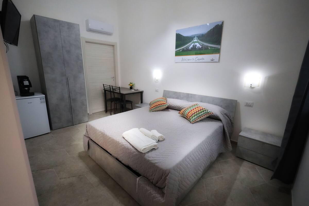 A bedroom with a bed, pillows, a desk, a chair, and a coffee machine.