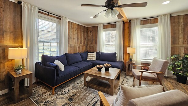 A modern living room with a dark blue sectional sofa, wooden accents, a ceiling fan, and large windows with blinds. - Charming Farmhouse in Oneonta, NY, 2 Mins away from Cooperstown All Star Village (Oneonta)