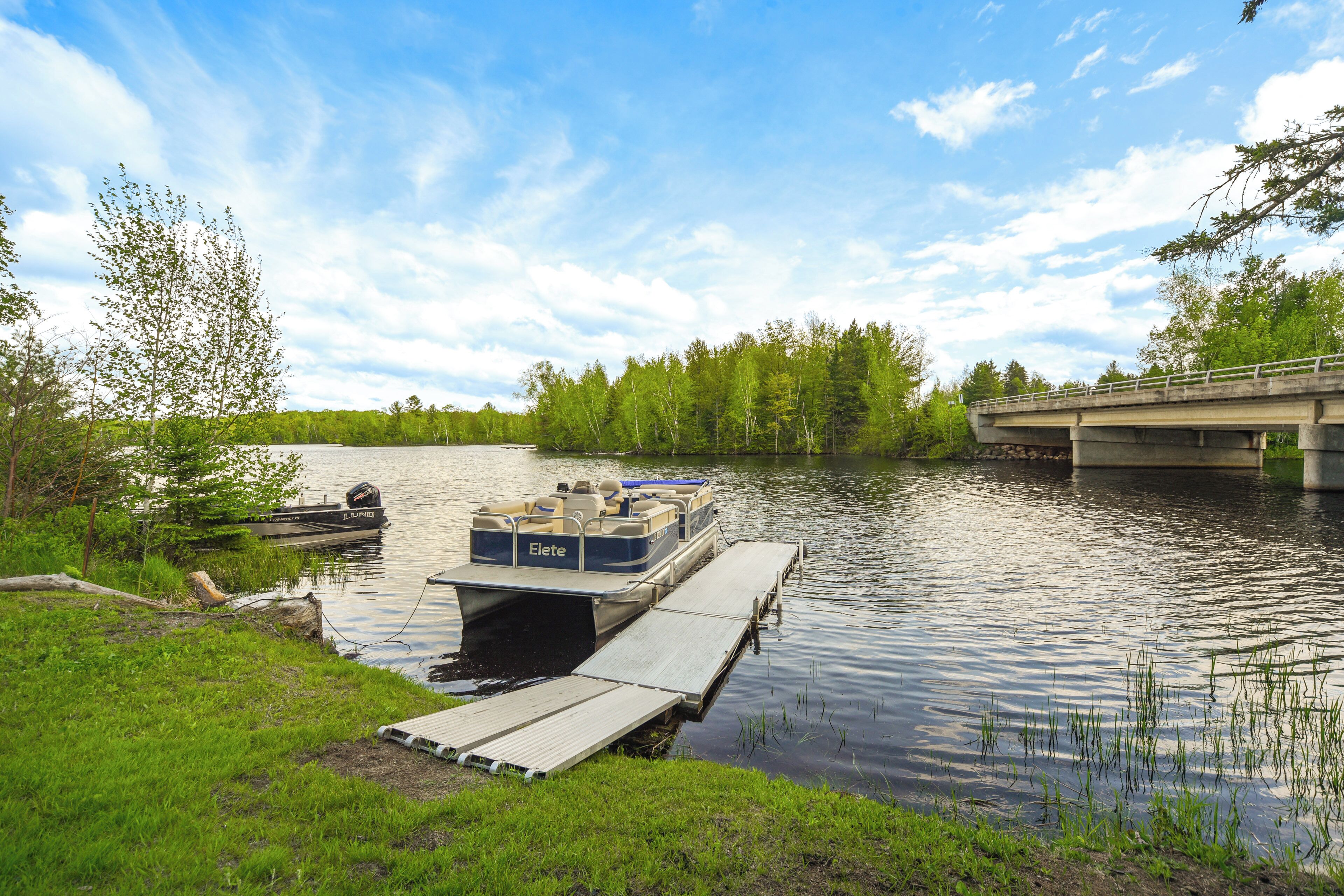 A boat docked at a wooden pier by a calm river, with a bridge in the background and trees lining the riverbank.