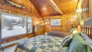 A wooden cabin interior with a large window, a bed with a quilt, a dresser, and a fish mounted on the wall. - Hayward Retreat on the Chippewa Flowage w/ Dock! (Hayward)