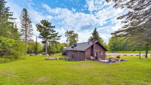 A cabin with a deck and outdoor seating area, surrounded by a grassy field and trees. - Hayward Retreat on the Chippewa Flowage w/ Dock! (Hayward)