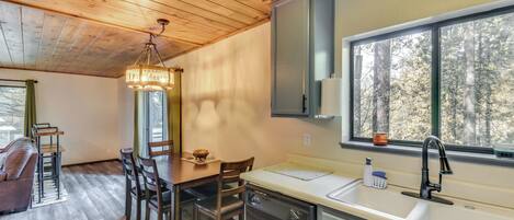A modern kitchen with wooden flooring, a dining area with a table and chairs, and a window with a view of trees.