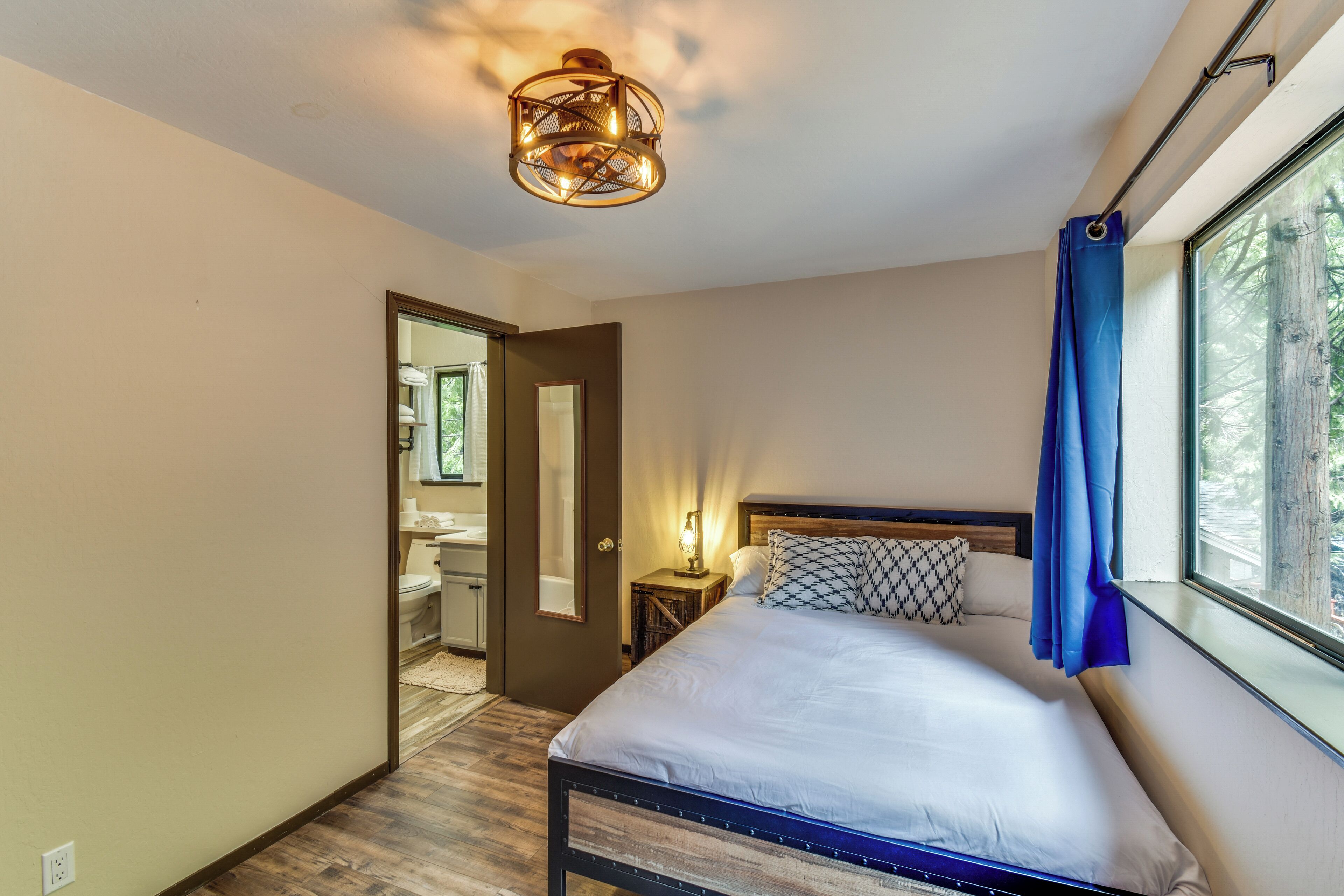 A bedroom with a bed, a nightstand, a mirror, a window with curtains, and a ceiling light fixture.