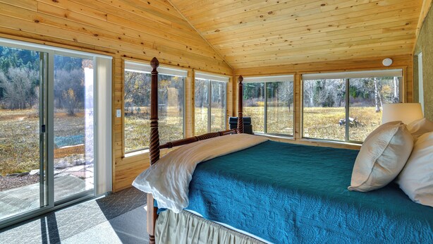 Ein Schlafzimmer einer Holzhütte mit einem großen Bett, einer Holzbalkendecke und großen Fenstern mit Blick ins Freie.