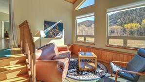 Ein gemütliches Wohnzimmer mit einer braunen Ledersofa, einem Holztisch und Blick auf einen bewaldeten Hang durch große Fenster.