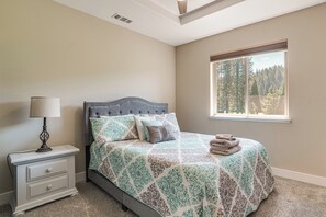 Ein Schlafzimmer mit einem Bett, einem Nachttisch, einer Lampe und einem Fenster mit Blick auf Bäume.