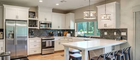 Una cocina moderna con armarios blancos, electrodomésticos de acero inoxidable, una isla central con taburetes altos e iluminación colgante.