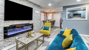 Ein modernes Wohnzimmer mit Steinwand, Flachbildfernseher, Kamin, einem gelben Sessel, einem blauen Sofa und einer Küchenzeile im Hintergrund.