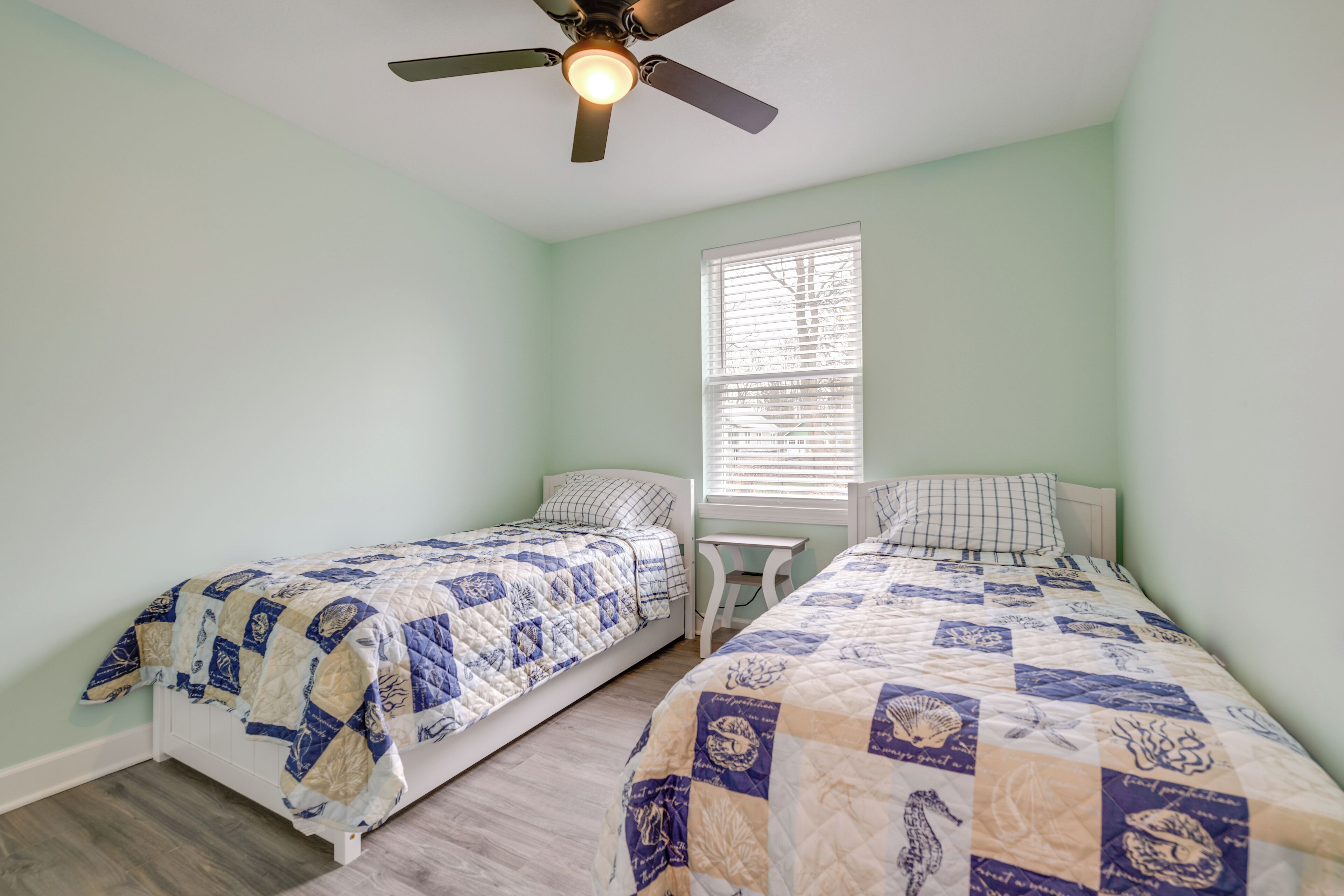 A bedroom with two beds, a ceiling fan, a window with blinds, and a small table.