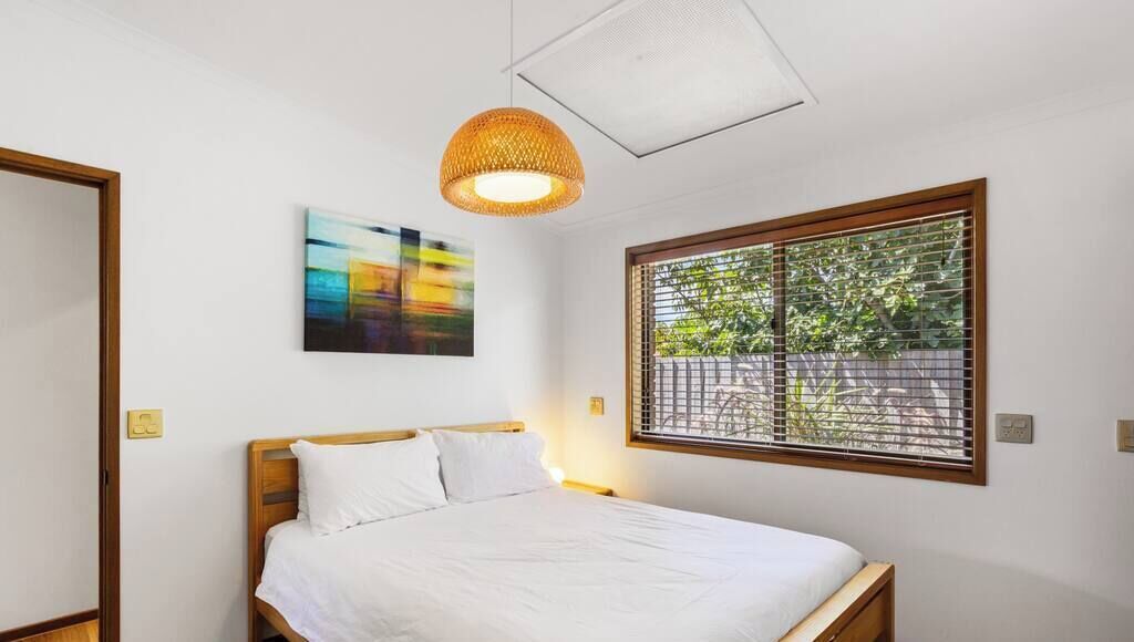 A bedroom with a bed, a hanging light fixture, a window with blinds, and a painting on the wall.
