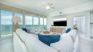 A spacious living room with a large white sofa, a coffee table, and a TV mounted on the wall. There is a balcony with a view of the ocean.