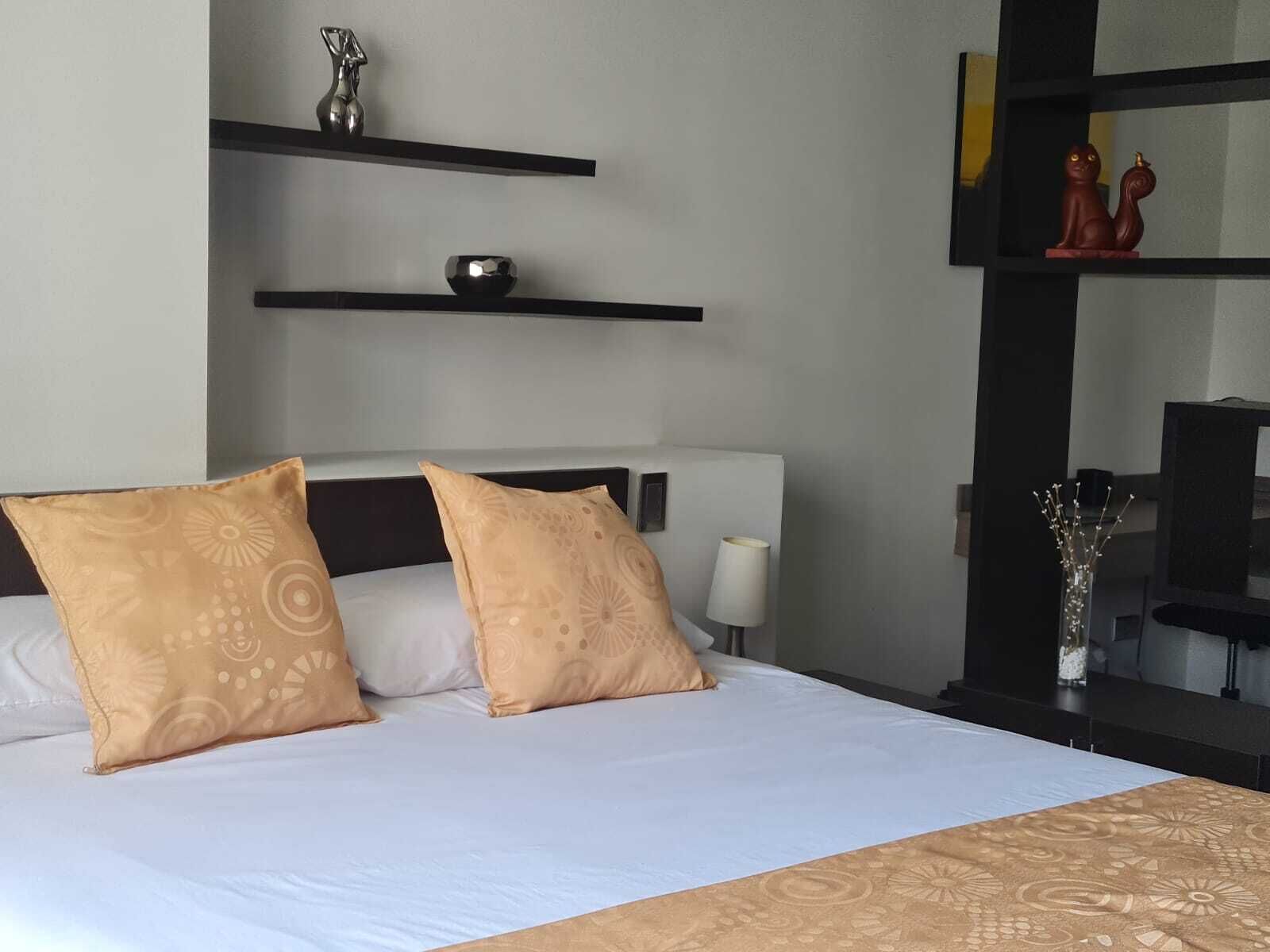 A hotel room with a bed, two decorative pillows, a bedside table with a lamp, and a shelf with decorative items.