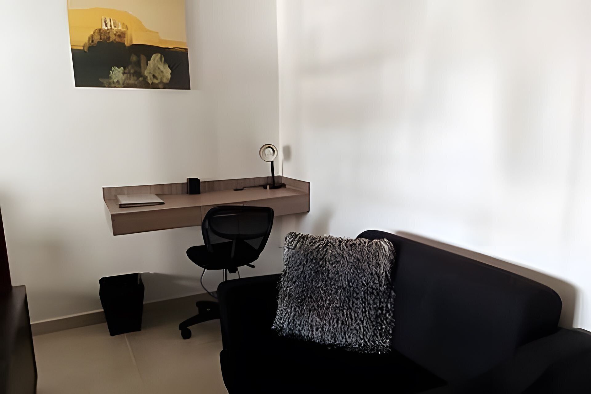 A modern living room with a black sofa, a small black office chair, a wooden desk with a computer, and a wall-mounted shelf.