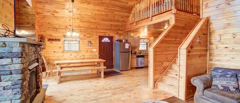 A wooden cabin interior with a stone fireplace, wooden benches, a sofa, a dining table, and a kitchen area.