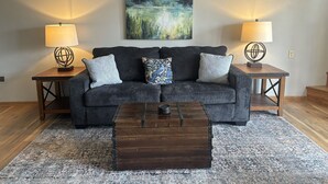 A living room with a dark grey sofa, two matching side tables, a wooden coffee table, and a large abstract painting on the wall. - The Bungalow on Lake Navajo (Cherokee Village)