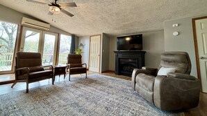 A living room with a fireplace, ceiling fan, and large windows. - The Bungalow on Lake Navajo (Cherokee Village)