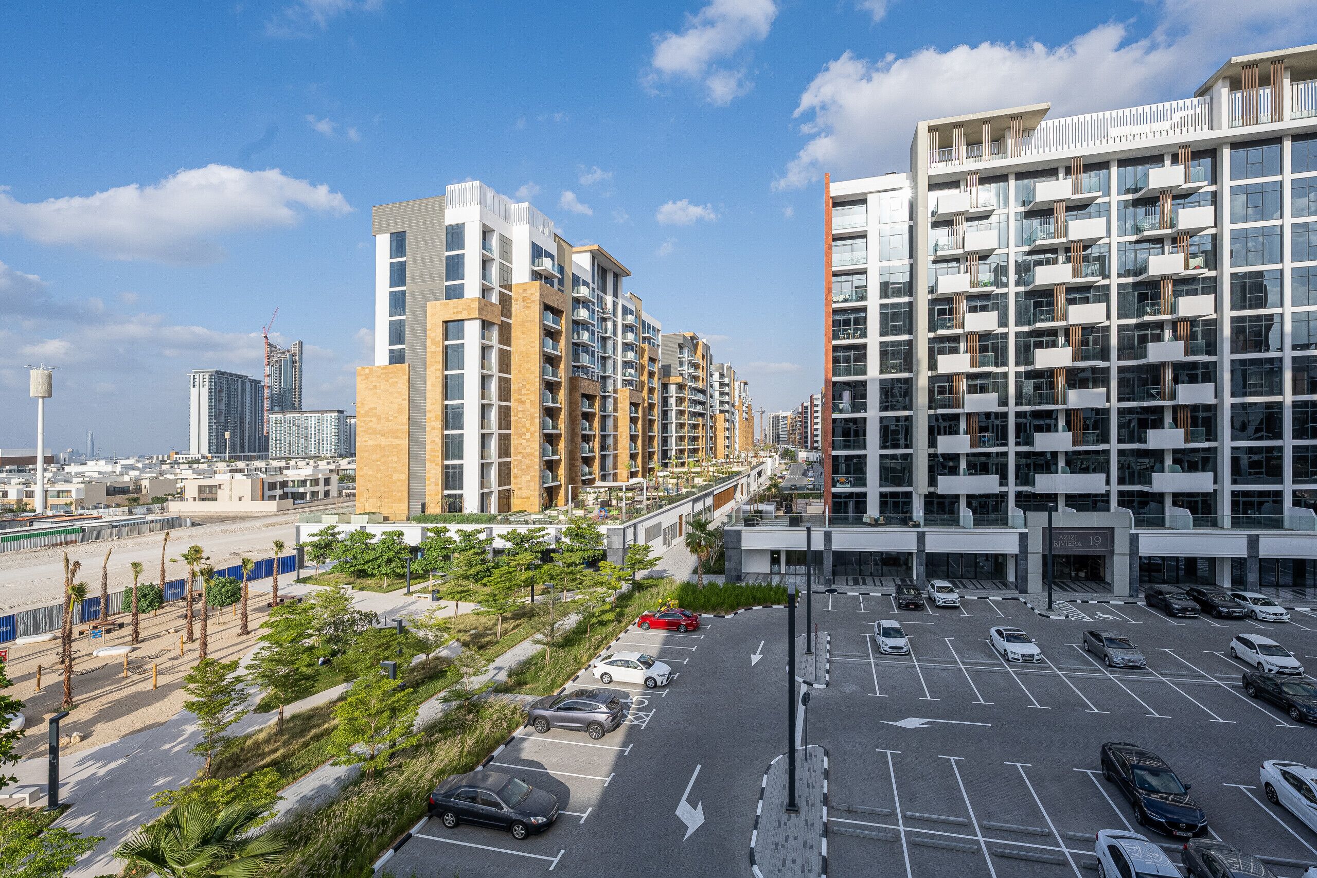 Un complejo moderno de apartamentos con varios edificios altos, un estacionamiento y un paseo marítimo frente a la playa.