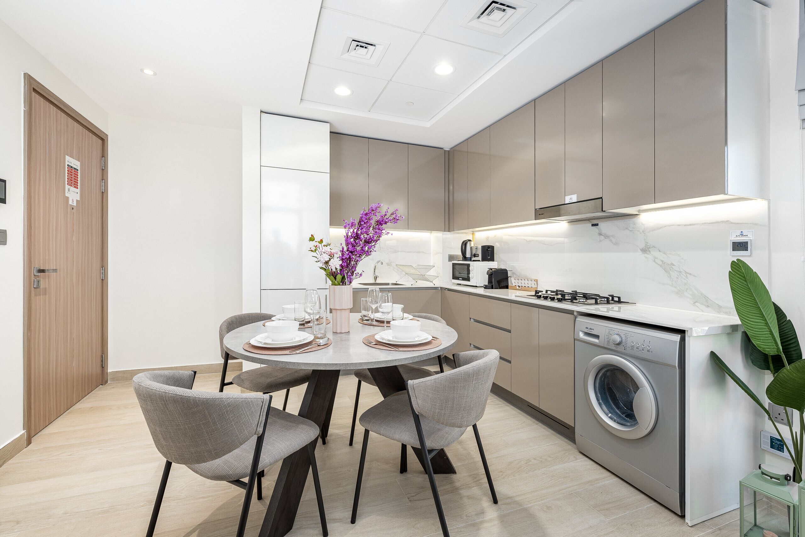 A modern kitchen with a built-in washing machine, a dining area with a round table and chairs, and a small plant on the counter.