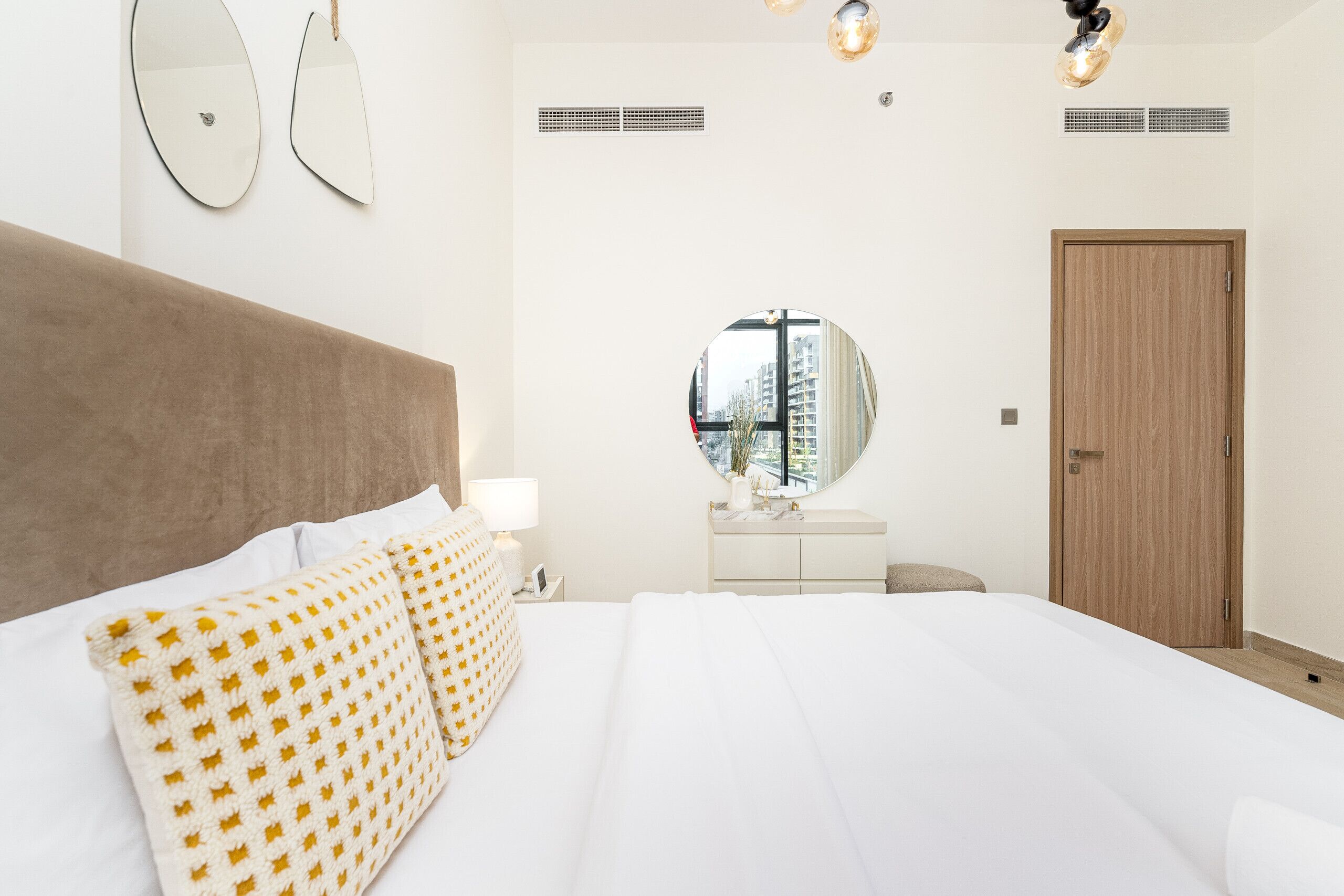 Habitación de hotel con una cama grande, un espejo redondo, una puerta de madera y vistas a edificios por la ventana.