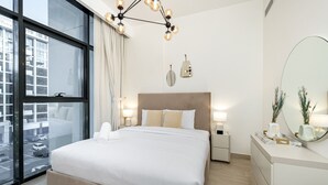 A modern hotel room with a large bed, a bedside table, a dresser, and a large window offering a city view. - Comfy 1BR Apartment in Azizi Riviera 8, Al Merkadh by Deluxe Holiday Homes (Dubai)