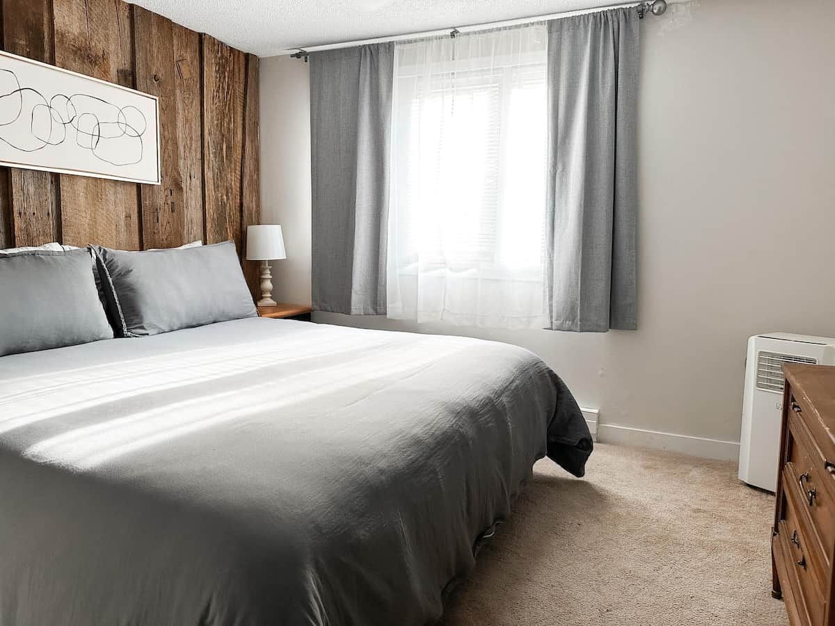 A bedroom with a bed, a nightstand, a lamp, a dresser, and a window with curtains.
