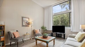 A living room with a sofa, armchair, coffee table, TV, bookshelf, and a window with curtains. - 1164 Beach Walker - Adorable oceanfront, just steps to the beach and pool! (Fernandina Beach)