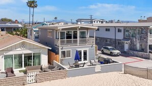 Un immeuble résidentiel de deux étages doté d’un balcon, avec une voiture garée devant et offrant une vue dégagée sur la plage.