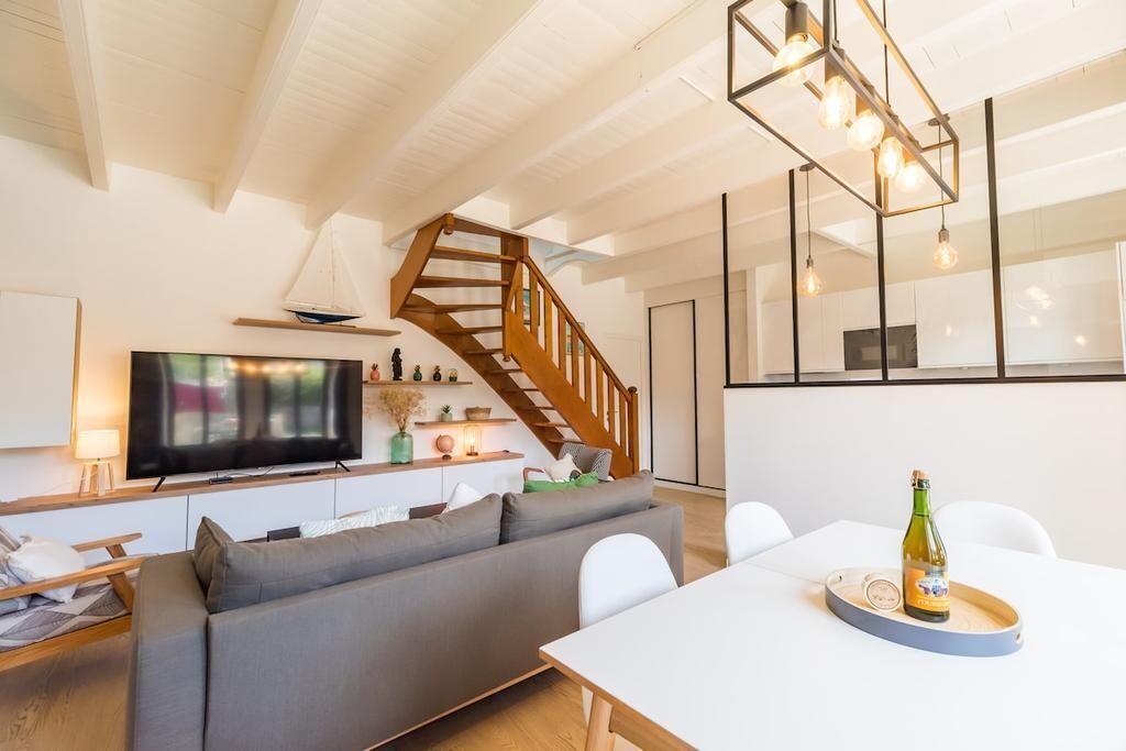 A modern living room with a grey sofa, a wooden staircase, a flat-screen TV, and a dining area with a table and chairs.