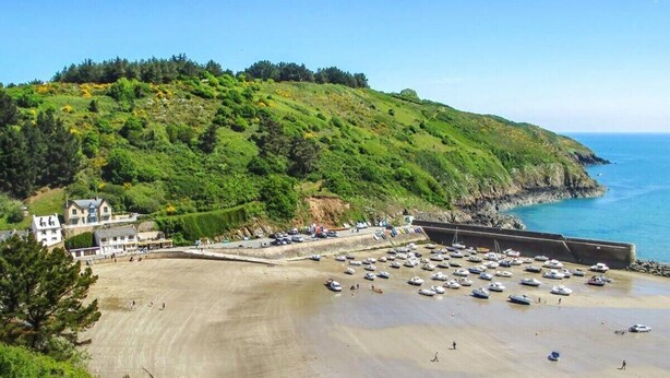 Une plage côtière avec un espace de stationnement, un bâtiment doté de nombreuses fenêtres et un paysage vallonné en arrière-plan.