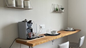 A kitchenette with a coffee maker, a shelf with jars, a small plant, and a dining area with a chair and table. - Pleasant 2-room apartment close to shops and train (Saint-Brieuc)