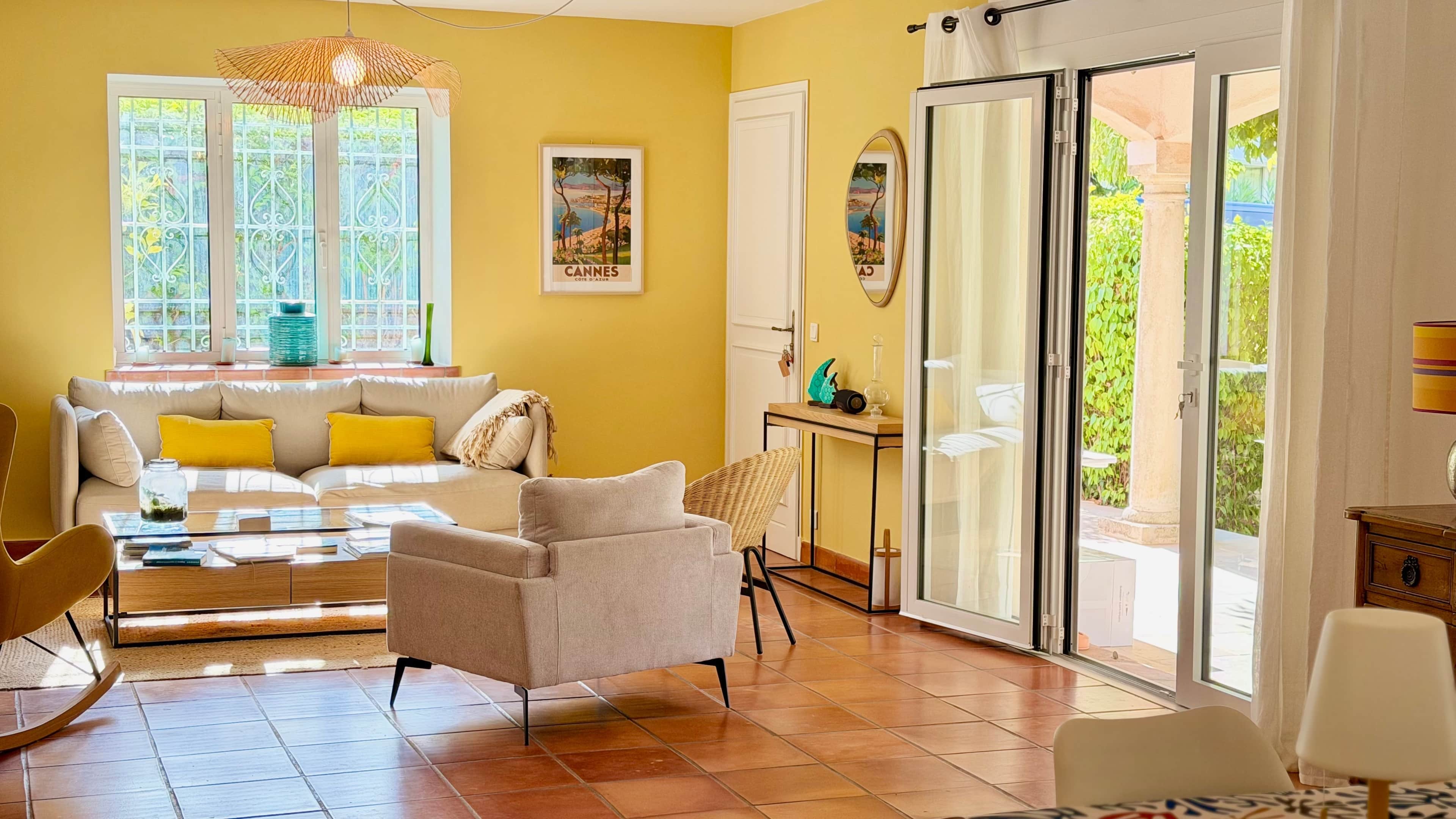 A bright living room with a sofa, armchairs, a glass door leading to a garden, and a window with decorative items.